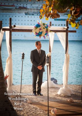 Officiant stands under a wedding arch as a cruise ship passes in the background